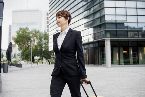 Weiblicher Unternehmer mit Koffer zu Fuß auf der Straße gegen moderne Gebäude in der Stadt - MEUF01272