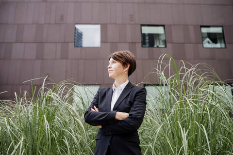 Thoughtful businesswoman with arms crossed standing by plants against building in city stock photo