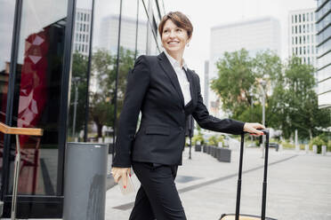 Smiling female entrepreneur with suitcase walking on city street - MEUF01287