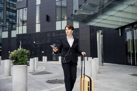 Female professional listening music while walking with suitcase against modern building stock photo