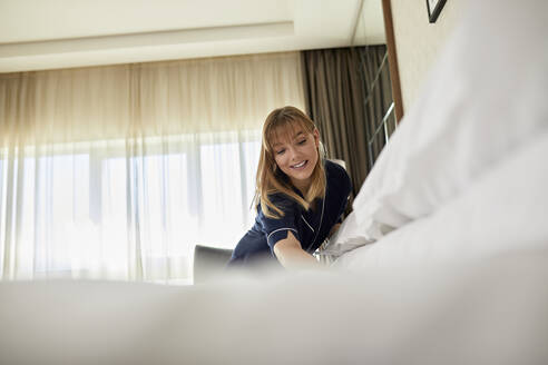 Lächelndes Zimmermädchen, das im Hotelzimmer stehend das Bett macht - ZEDF03472