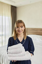 Lächelndes Zimmermädchen mit Handtüchern in der Hand im Schlafzimmer eines Hotels - ZEDF03510