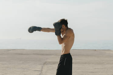 Shirtless young man practicing boxing while standing at harbor on sunny day - VABF03108