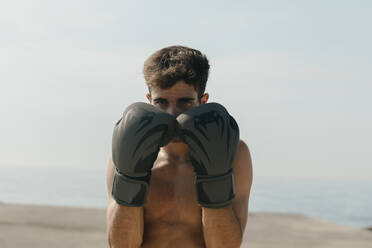 Nahaufnahme eines hemdsärmeligen Boxers mit Handschuhen, der am Hafen vor einem klaren Himmel steht - VABF03109