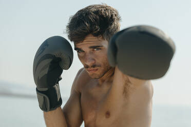 Close-up of shirtless boxer wearing gloves practicing boxing against clear sky - VABF03111