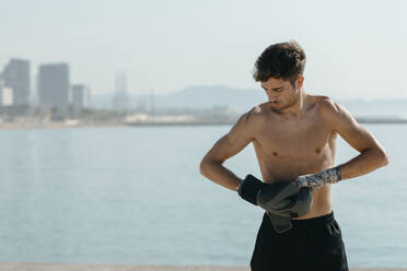 Shirtless boxer wearing gloves while standing against sea on sunny day - VABF03112