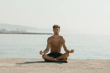 Shirtless young man meditating while sitting against sea at harbor - VABF03113