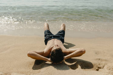 Shirtless young man with hands behind head lying on sand at beach during sunny day - VABF03115