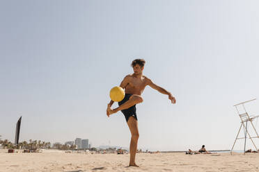 Shirtless male athlete playing with ball at beach against clear sky on sunny day - VABF03117