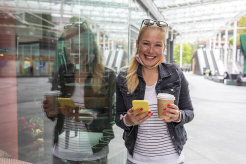 Lächelnde Frau, die eine Kaffeetasse hält und ein Smartphone benutzt, während sie auf dem Gehweg in der Stadt steht - WPEF03163