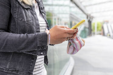 Nahaufnahme einer mittleren erwachsenen Frau, die eine Maske hält und ein Smartphone benutzt, während sie im Freien steht - WPEF03164