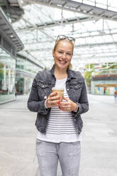 Smiling mid adult woman holding coffee cup while standing on sidewalk in city - WPEF03165