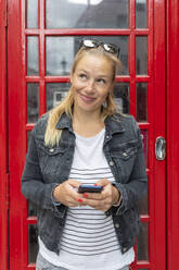 Lächelnde Frau, die ein Smartphone hält und wegschaut, während sie an einer Telefonzelle steht - WPEF03168
