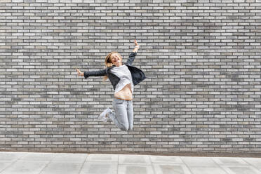 Carefree woman with arms outstretched jumping on footpath against gray brick wall - WPEF03170