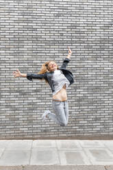Glückliche Frau mit ausgestreckten Armen, die auf dem Fußweg gegen eine graue Backsteinmauer springt - WPEF03171
