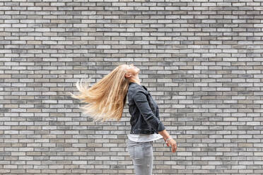 Woman tossing long hair while standing by gray brick wall - WPEF03173