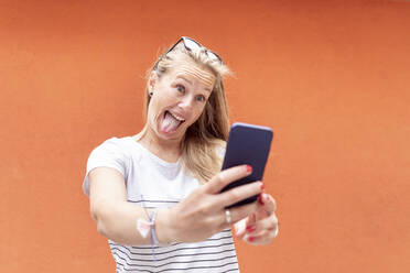 Playful woman sticking out tongue while taking selfie with smart phone against orange wall - WPEF03177