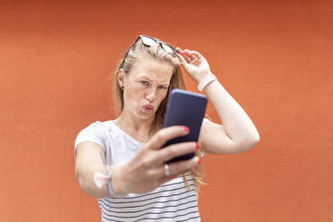 Frau spitzt die Lippen, während sie ein Selfie mit ihrem Smartphone an einer orangefarbenen Wand macht - WPEF03178