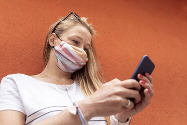 Woman wearing mask using smart phone while standing against orange wall - WPEF03179