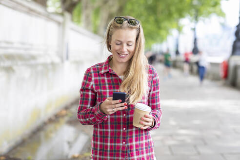Smiling mid adult woman holding coffee using mobile phone while standing on footpath - WPEF03182