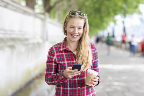 Smiling woman holding coffee using mobile phone while standing on footpath - WPEF03183