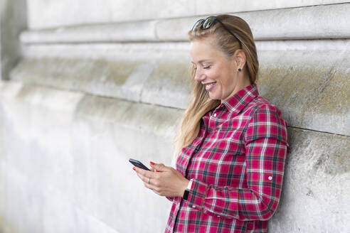 Lächelnde Frau mit kariertem Hemd, die ein Mobiltelefon benutzt, während sie sich an die Wand lehnt - WPEF03184