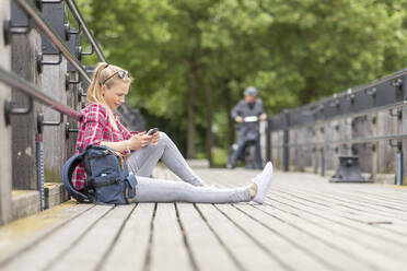 Mittlere erwachsene Frau, die ein Smartphone benutzt, während sie auf einer Brücke in der Stadt sitzt - WPEF03188