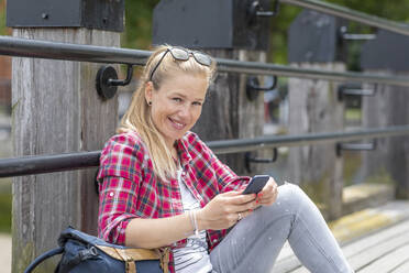 Lächelnde Frau im mittleren Erwachsenenalter, die ein Smartphone benutzt, während sie an der Reling auf dem Pier sitzt - WPEF03190