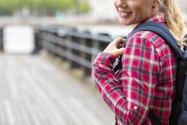 Close-up of smiling woman wearing checked shirt carrying backpack while standing on bridge - WPEF03199