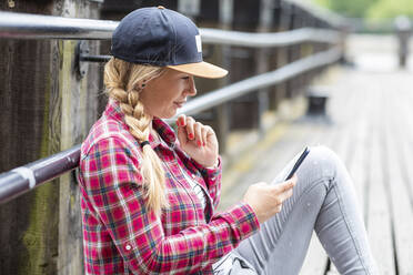 Frau mit geflochtenem Haar, die auf einer Brücke sitzend ein Smartphone benutzt - WPEF03204