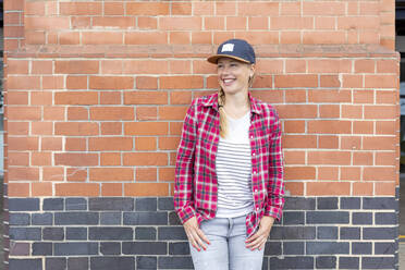 Lächelnde Frau mit kariertem Hemd und Mütze, die an einer Backsteinmauer steht - WPEF03206