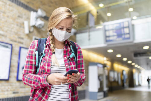 Woman wearing mask using smart phone while standing at tube station - WPEF03209