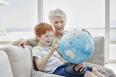 Großmutter und Enkel sitzen auf einer Couch in einer Villa und betrachten einen Globus - RORF02249