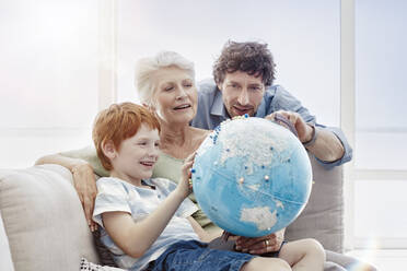 Großmutter, Vater und Enkel sitzen auf einer Couch in einer Villa und betrachten einen Globus - RORF02251