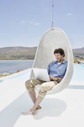 Man sitting in hanging chair above swimming pool using laptop - RORF02274