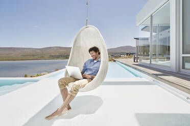 Man sitting in hanging chair above swimming pool using laptop - RORF02277