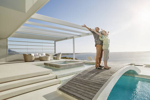 Unbeschwertes älteres Paar, das auf einer Brücke über einem Swimmingpool in einem luxuriösen Strandhaus steht, lizenzfreies Stockfoto