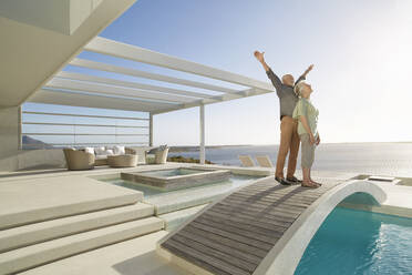 Unbeschwertes älteres Paar, das auf einer Brücke über einem Swimmingpool in einem luxuriösen Strandhaus steht - RORF02310