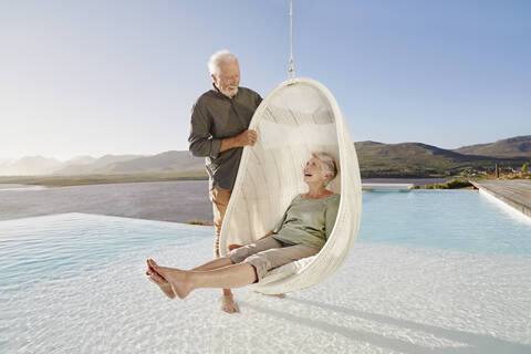 Glückliches älteres Paar mit Frau, die in einem Hängesessel über dem Schwimmbad sitzt, lizenzfreies Stockfoto