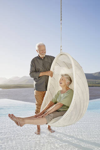 Glückliches älteres Paar mit Frau, die in einem Hängesessel über dem Schwimmbad sitzt, lizenzfreies Stockfoto