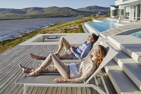 Couple lying on deck chairs at luxury beach house - RORF02323