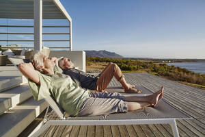 Senior couple lying on deck chairs at luxury beach house - RORF02329