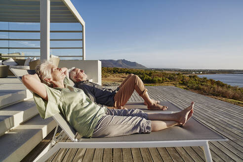 Senior couple lying on deck chairs at luxury beach house - RORF02329