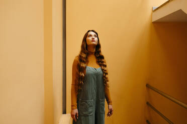 Junge Frau im Overall, die sich an eine orangefarbene Wand lehnt und nach oben schaut - TCEF00900