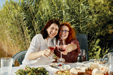 Porträt von zwei glücklichen Freundinnen beim Abendessen am See, die ihre Weingläser erheben, lizenzfreies Stockfoto