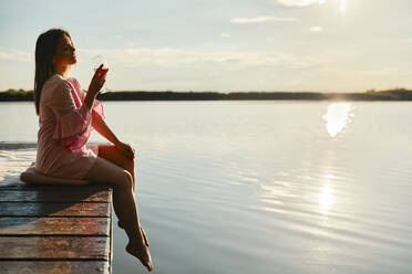 Junge Frau mit einem Glas Wein auf einem Steg an einem See bei Sonnenuntergang sitzend - ZEDF03578