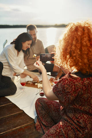Frau nimmt Smartphone Foto von Freunden mit Picknick auf dem Steg an einem See bei Sonnenuntergang, lizenzfreies Stockfoto