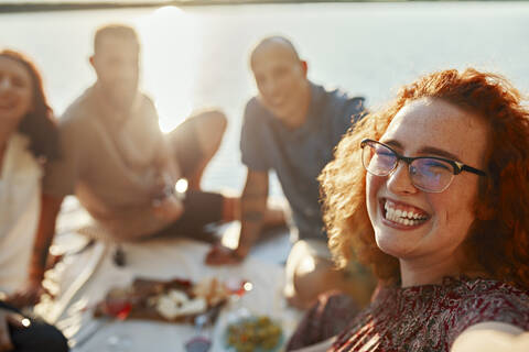 Glückliche rothaarige Frau beim Picknick mit Freunden auf einem Steg an einem See bei Sonnenuntergang, lizenzfreies Stockfoto