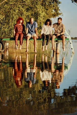 Freunde, die sich im Wasser spiegeln, sitzen auf einem Steg an einem See, lizenzfreies Stockfoto
