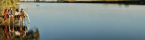 Panoramablick auf Freunde, die sich im Wasser spiegeln und auf einem Steg an einem See sitzen, lizenzfreies Stockfoto